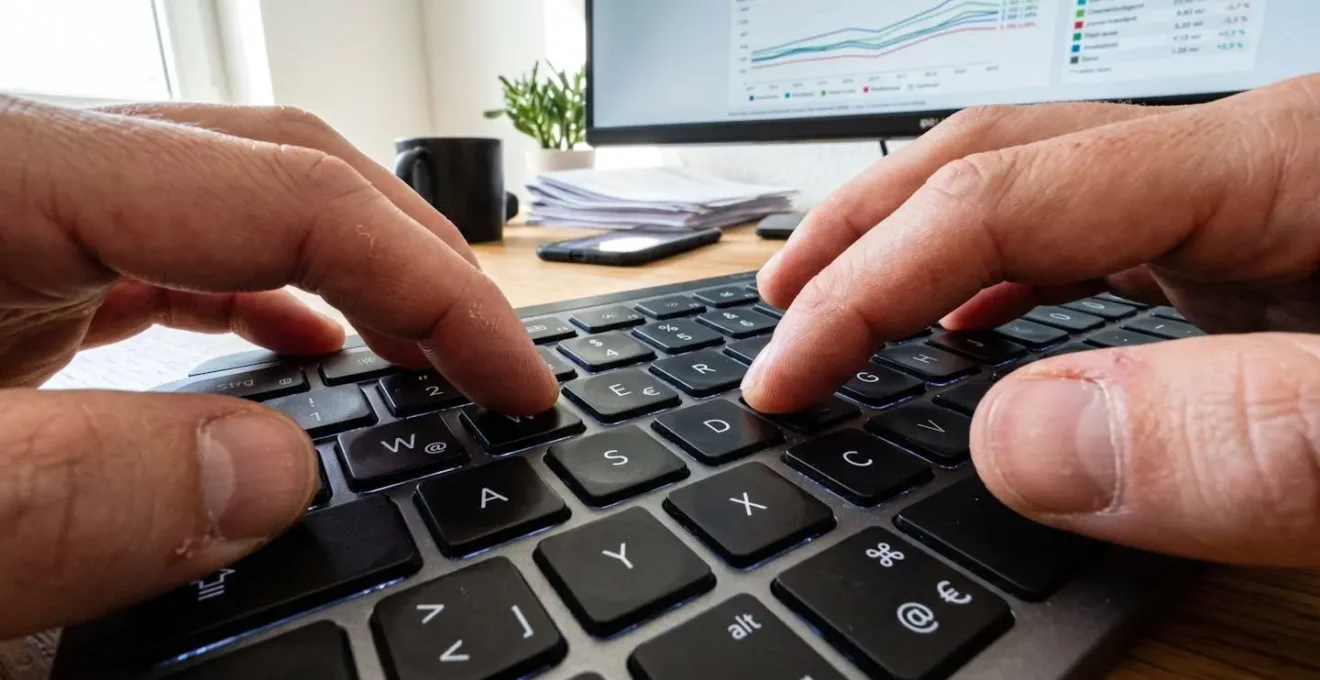 Extreme Nahaufnahme von Fingern, die auf einer modernen Tastatur tippen, im unscharfen Hintergrund ein Bildschirm mit verschwommenen Immobiliengrafiken